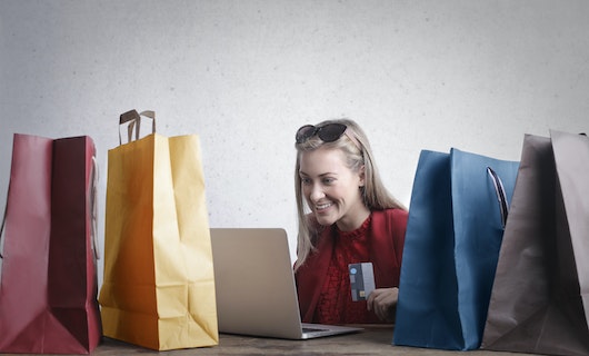 Junge Frau sitzt zwischen Einkaufstüten vor dem Laptop