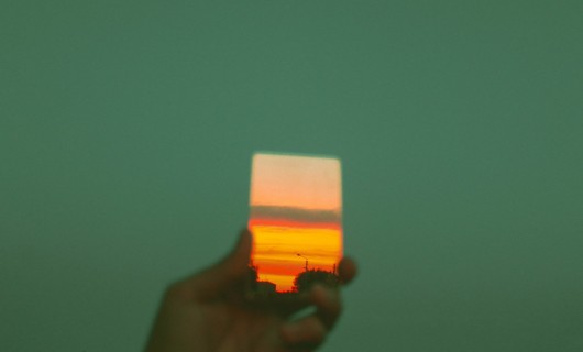 Vor einem grünen Hintergrund hält eine Hand ein Bild hoch auf dem ein Sonnenuntergang zu sehen ist.