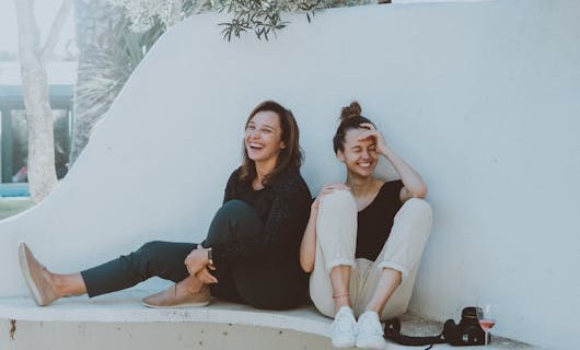 Zwei junge Mädchen sitzend lachend vor einer weißen Wand